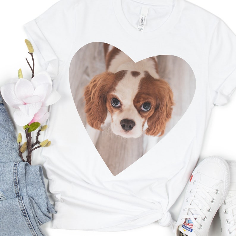 May include: A white short-sleeved t-shirt featuring a large heart-shaped graphic on the chest. Inside the heart is a photographic print of a brown and white Cavalier King Charles Spaniel looking upward with large, expressive eyes and floppy ears.