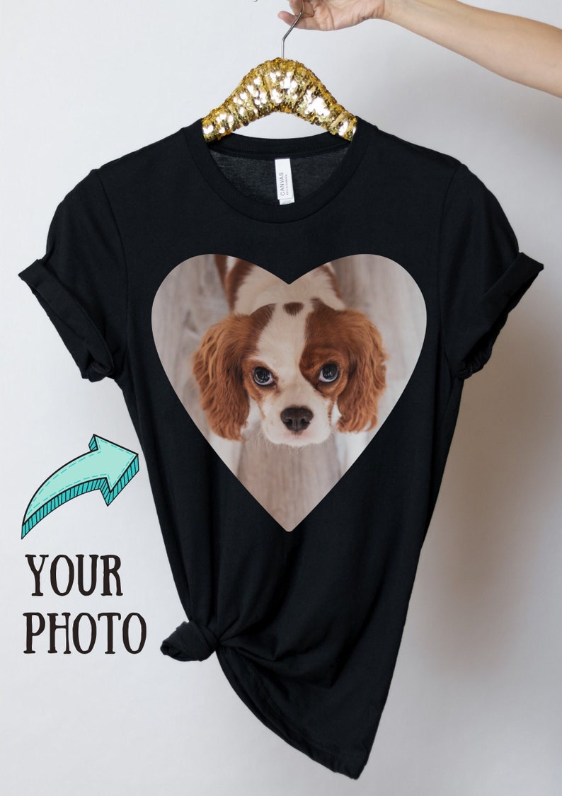 May include: A black short-sleeved t-shirt featuring a heart-shaped graphic containing a photograph of a brown and white Cavalier King Charles Spaniel. To the left of the heart graphic, there is a light blue arrow pointing toward the text YOUR PHOTO.