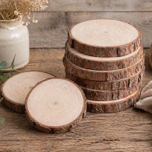 May include: A stack of rustic wooden coasters with natural bark edges. Two additional coasters are placed nearby. The coasters are made of light-colored wood and are arranged on a wooden surface. Decorative elements include a vase with dried flowers and a ball of twine.