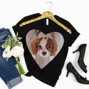 May include: A black t-shirt featuring a heart-shaped graphic of a brown and white Cavalier King Charles Spaniel dog. The shirt is displayed with blue denim jeans, a bouquet of white flowers, and a pair of black high-heeled shoes.