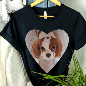 May include: A black short-sleeved t-shirt featuring a large heart-shaped graphic on the chest. Inside the heart is a photographic print of a brown and white Cavalier King Charles Spaniel with long ears and large dark eyes looking upward.