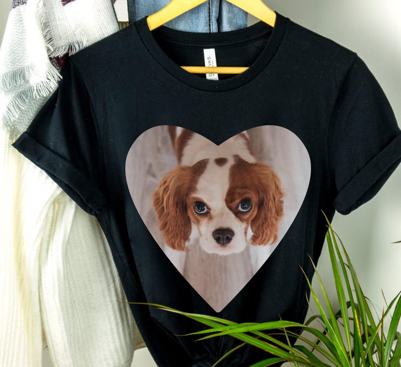 May include: A black short-sleeved t-shirt featuring a large heart-shaped graphic on the chest. Inside the heart is a photographic print of a brown and white Cavalier King Charles Spaniel with long ears and large dark eyes looking upward.