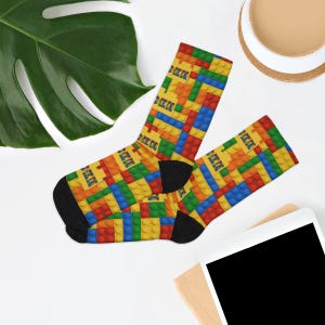 K&ouml;nnte beinhalten: Ein Paar Socken mit einem lebendigen, sich wiederholenden Muster aus ineinandergreifenden Kunststoff-Bausteinen in Prim&auml;rfarben wie Rot, Blau, Gelb und Gr&uuml;n. Die Socken haben einfarbig schwarze Fersen und Zehenbereiche, die einen deutlichen Kontrast zum bunten Blockdesign bilden.