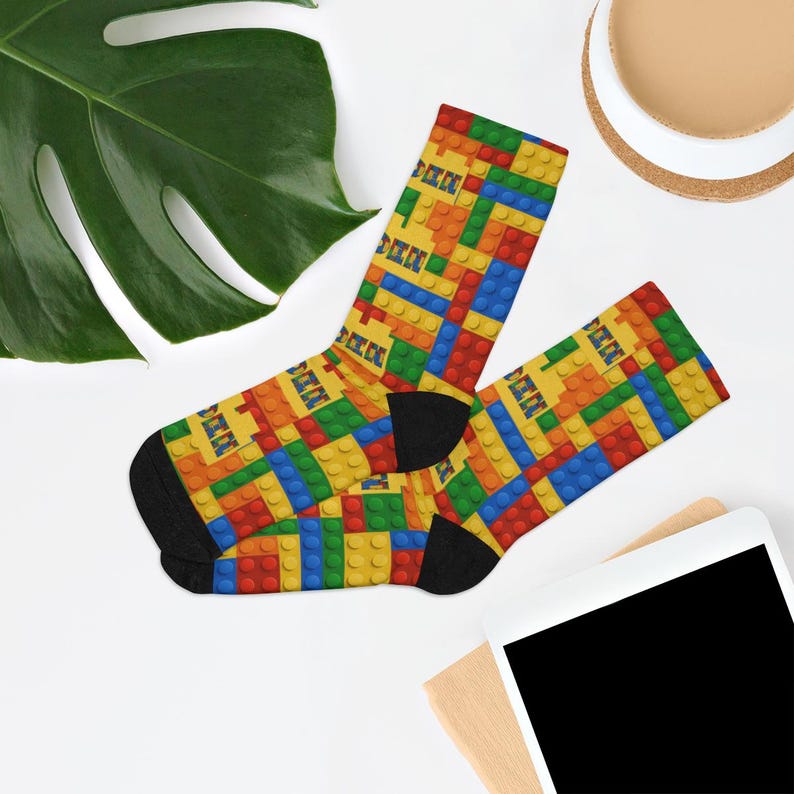 K&ouml;nnte beinhalten: Ein Paar Socken mit einem lebendigen, sich wiederholenden Muster aus ineinandergreifenden Kunststoff-Bausteinen in Prim&auml;rfarben wie Rot, Blau, Gelb und Gr&uuml;n. Die Socken haben einfarbig schwarze Fersen und Zehenbereiche, die einen deutlichen Kontrast zum bunten Blockdesign bilden.