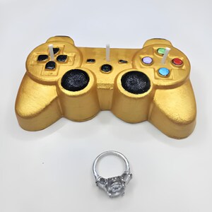 May include: A gold-colored video game controller with a silver ring on a white surface.