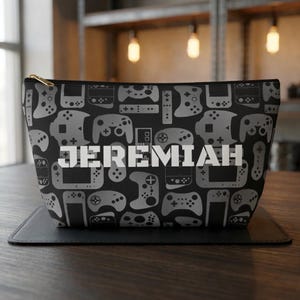 May include: Black cosmetic bag with a pattern of gray video game controllers. The name "JEREMIAH" is printed in large white letters across the center. The bag has a gold zipper and is sitting on a black surface.