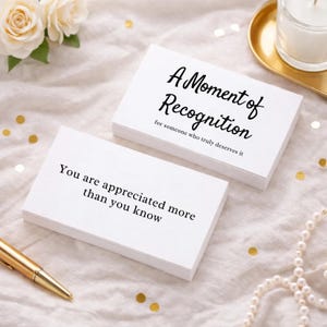 May include: White cards with black text, stacked and arranged on a soft, textured surface. The top card reads "A Moment of Recognition" and "for someone who truly deserves it." Another card says "You are appreciated more than you know."