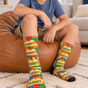 K&ouml;nnte beinhalten: Ein Paar wadenlange Socken mit einem bunten Muster aus ineinandergreifenden Kunststoff-Bausteinen in Rot, Gelb, Blau und Gr&uuml;n. Auf jeder Socke ist der Name GRANT in fetten blauen Gro&szlig;buchstaben auf einem gelben rechteckigen Block gedruckt.