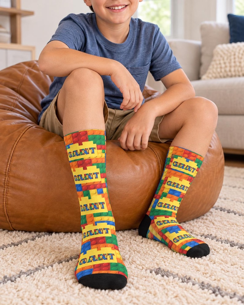 K&ouml;nnte beinhalten: Ein Paar wadenlange Socken mit einem bunten Muster aus ineinandergreifenden Kunststoff-Bausteinen in Rot, Gelb, Blau und Gr&uuml;n. Auf jeder Socke ist der Name GRANT in fetten blauen Gro&szlig;buchstaben auf einem gelben rechteckigen Block gedruckt.