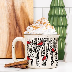 Puede incluir: Taza de cerámica blanca con una escena de invierno de abedules y cardenales rojos. La taza tiene un esmalte moteado y está llena de crema batida blanca.