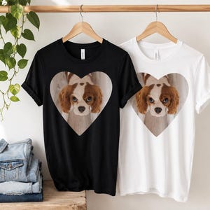 May include: A black t-shirt and a white t-shirt, each featuring a central heart-shaped graphic containing a photographic print of a brown and white Cavalier King Charles Spaniel dog looking directly forward with large, expressive eyes.