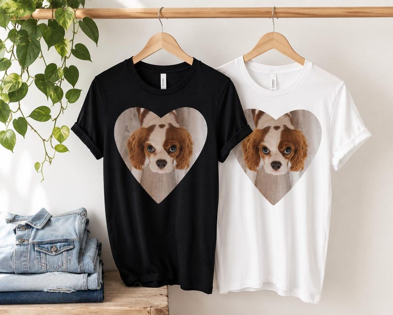 May include: A black t-shirt and a white t-shirt, each featuring a central heart-shaped graphic containing a photographic print of a brown and white Cavalier King Charles Spaniel dog looking directly forward with large, expressive eyes.