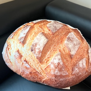 May include: A large, round, brown and white bread cushion. The cushion is shaped like a loaf of bread and has a realistic texture.