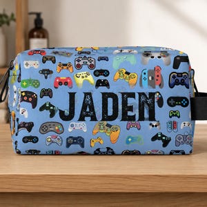 May include: A blue rectangular cosmetic bag with a pattern of colorful video game controllers. The name "JADEN" is printed in large black letters across the center. The bag has a black handle on the side.