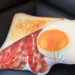 May include: A white pillow shaped like a slice of toast with a fried egg and bacon on top. The toast is golden brown, the egg yolk is bright yellow, and the bacon is red and crispy.
