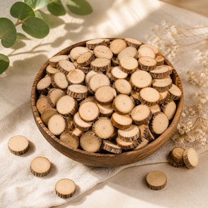 Peut inclure: Un bol en bois rempli de nombreuses petites tranches de bois rondes. Les tranches sont brun clair avec un grain de bois et des bords d'écorce visibles. Plusieurs tranches sont éparpillées autour du bol sur un tissu de couleur neutre. Fournitures d'artisanat.