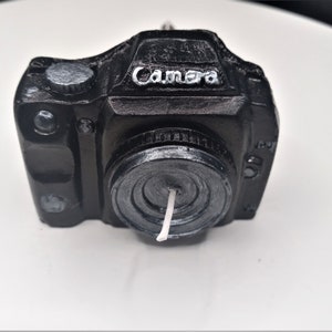 May include: Black camera shaped candle with a white wick. The candle has the word "Camera" written on the top.