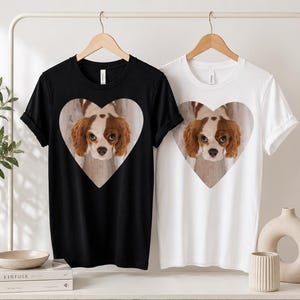 May include: A black t-shirt and a white t-shirt, each featuring a central heart-shaped graphic containing a photographic print of a brown and white Cavalier King Charles Spaniel dog with large eyes looking directly forward at the viewer.