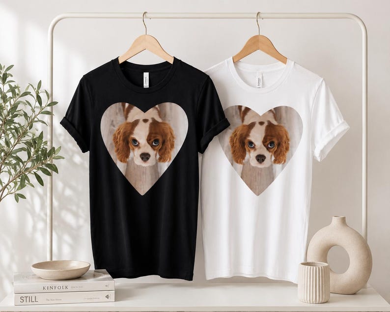 May include: A black t-shirt and a white t-shirt, each featuring a central heart-shaped graphic containing a photographic print of a brown and white Cavalier King Charles Spaniel dog with large eyes looking directly forward at the viewer.