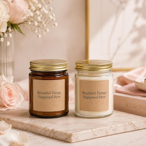 May include: Two glass jar candles with gold lids, one brown and one clear, sit on a marble surface. Each candle has a brown label with the text "Beautiful Things Happened Here". A bouquet of pink roses and a book are in the background.