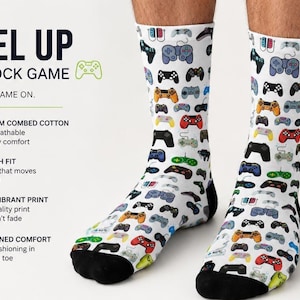 May include: White crew socks featuring a repeating pattern of colorful video game controllers in various styles. The socks have solid black heels and toes. Text on the left reads: LEVEL UP YOUR SOCK GAME, COMFY. FUN. GAME ON. PREMIUM COMBED COTTON.