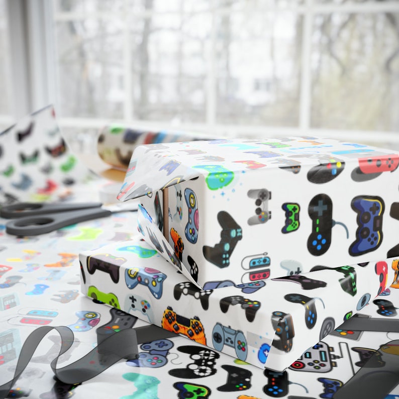 May include: Two gift boxes wrapped in white paper with colourful video game controller designs. The wrapping paper is decorated with various game controller illustrations in shades of blue, green, orange, and black. Scissors and a roll of matching paper are visible.