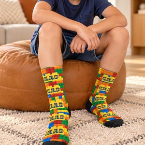 May include: A pair of crew socks featuring a colorful pattern of interlocking plastic building blocks in red, yellow, blue, and green. The word CHAD is printed in blue capital letters repeatedly across the block design. The socks have black toes.