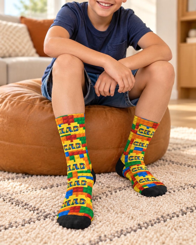 K&ouml;nnte beinhalten: Ein Paar Socken mit einem bunten Muster aus ineinandergreifenden Kunststoff-Bausteinen in Rot, Gelb, Blau und Gr&uuml;n. Das Wort CHAD ist wiederholt in blauen Gro&szlig;buchstaben &uuml;ber das Blockdesign gedruckt. Die Socken haben schwarze Zehenbereiche.