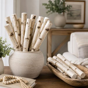 May include: A collection of birch logs with natural bark patterns, displayed in a cream-colored ceramic vase and a wooden bowl. The logs have a light beige and brown color scheme, creating a rustic, natural aesthetic. Decorative home decor.
