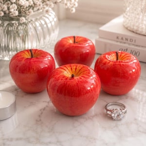 May include: Four glossy red decorative apples with yellow-orange gradients and thin brown stems. Each apple features a smooth, reflective surface. A silver-toned ring with a clear, faceted stone is positioned near the front right apple.