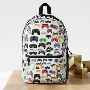 May include: White backpack covered in colorful illustrations of video game controllers. The backpack has a black top handle, a front pocket, and a black zipper. The controllers are in various colors and designs.
