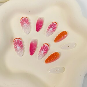 May include: A collection of artificial nails in various designs. Some nails feature a pink and white citrus fruit slice design with an orange rind. Other nails are solid pink or orange, and some are white with a gold dot.