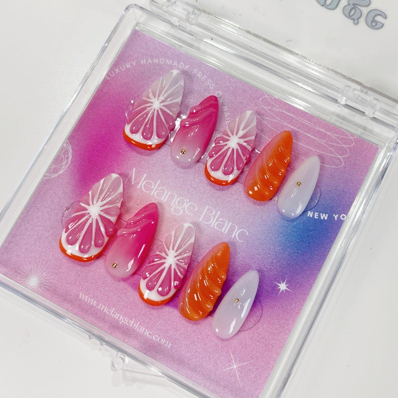 May include: A set of press-on nails in a clear plastic case. The nails feature various designs, including pink citrus fruit slices, orange segments, and solid pink and lavender nails. The text "Melange Blanc" and "Luxury Handmade Press On Nails" are visible.