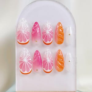 May include: A set of ten artificial nails with a fruit-inspired design. The nails feature a variety of shapes and colors, including pink, orange, and white. Some nails are designed to look like citrus fruit slices, while others have a swirl design.