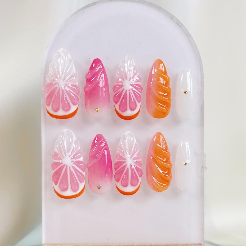 May include: A set of ten artificial nails with a fruit-inspired design. The nails feature a variety of shapes and colors, including pink, orange, and white. Some nails are designed to look like citrus fruit slices, while others have a swirl design.