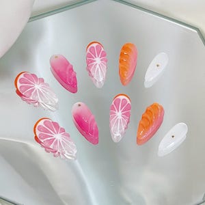 May include: A set of ten artificial nails with a fruit theme. The nails feature designs of pink and white citrus slices, orange bread, and solid pink and white nails with gold accents. The nails are arranged on a reflective surface.