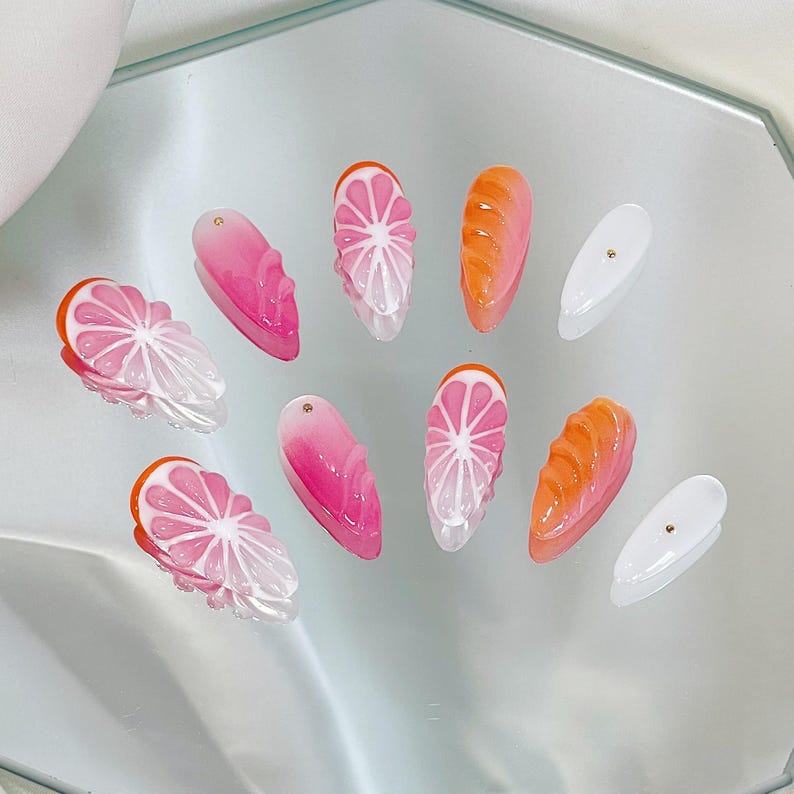 May include: A set of ten artificial nails with a fruit theme. The nails feature designs of pink and white citrus slices, orange bread, and solid pink and white nails with gold accents. The nails are arranged on a reflective surface.