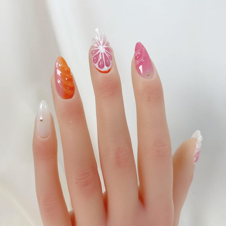 May include: A hand with long, almond-shaped nails featuring various designs. One nail has a white and pink citrus slice design, while others showcase orange and pink gradients and textured patterns. Each nail is adorned with a small gold stud.