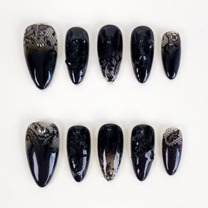 Puede incluir: Un conjunto de diez uñas postizas negras con acabado brillante. Algunas uñas presentan un delicado diseño de encaje negro, mientras que otras son de color negro liso. Las uñas de forma ovalada están dispuestas sobre una superficie blanca.
