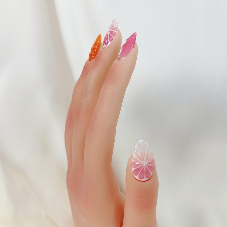 May include: A hand with long, almond-shaped nails. The nails feature a variety of designs, including a textured orange carrot, a pink and white citrus slice, and a gradient pink design. The nails are painted with gel polish.