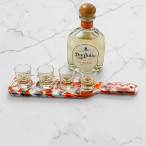 May include: A Don Julio Reposado tequila bottle with a wooden cap and a clear glass bottle body, accompanied by a rectangular serving paddle featuring a marbled orange, black, and white pattern, holding four clear glass shot glasses filled with liquid.