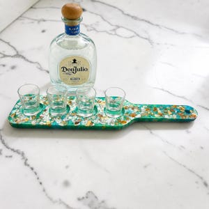 May include: A clear glass Don Julio Blanco tequila bottle with a wooden stopper and a rectangular resin serving paddle featuring a green and white marbled pattern with embedded shell fragments and four clear glass shot glasses.