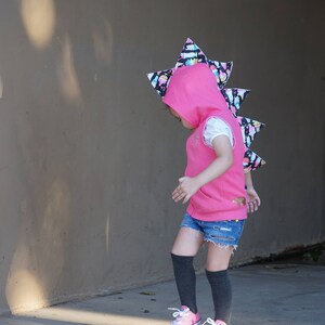 K&ouml;nnte beinhalten: Eine rosa Kapuzenweste mit einem schwarz-wei&szlig;en Dinosaurierschwanz. Die Weste hat einen Rei&szlig;verschluss und eine Kapuze mit einem schwarz-wei&szlig;en Dinosaurier-Design. Das Kind tr&auml;gt Jeans-Shorts und schwarze Kniestr&uuml;mpfe.