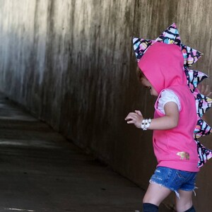 K&ouml;nnte beinhalten: Ein rosa Kapuzenpullover mit einem bunten Dinosaurierschwanz. Der Schwanz ist aus Stoff mit einem schwarz-wei&szlig;en Muster. Der Pullover hat ein goldenes B&auml;renlogo auf der Vorderseite.