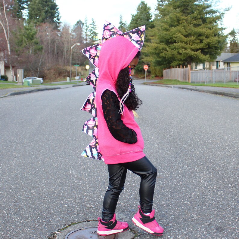 K&ouml;nnte beinhalten: Ein rosa Hoodie mit einem schwarzen und wei&szlig;en Dinosaurier-Schwanz-Design. Der Hoodie hat eine Kapuze und einen Rei&szlig;verschluss. Die Person tr&auml;gt eine schwarze Hose und rosa Schuhe.
