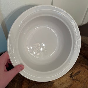May include: A white ceramic bowl with a wide rim and a slightly curved interior. The bowl is held by a hand and sits on a wooden surface. The bowl has a simple, clean design.