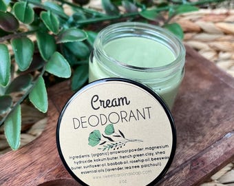 May include: A jar of cream deodorant with a light green hue, set on a wooden board with green foliage. The label reads "Cream DEODORANT" with a list of ingredients. The jar contains 2 oz of product.