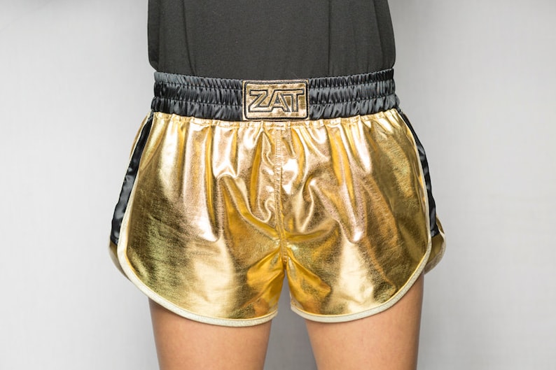 May include: Gold and black boxing shorts with a white trim and the word "ZAT" on the waistband.