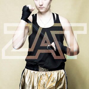 May include: A woman wearing black tank top with a gold belt and gold shorts with white trim. She is holding a black training stick in her hands. The word "ZAT" is written in white letters on the belt.