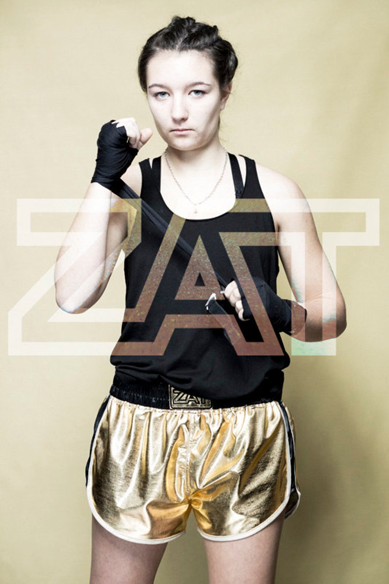 May include: A woman wearing black tank top with a gold belt and gold shorts with white trim. She is holding a black training stick in her hands. The word "ZAT" is written in white letters on the belt.